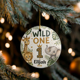 A personalized round ceramic Christmas ornament hanging from a pine tree branch. The ornament features safari animal illustrations including a giraffe, lion cub, and elephant, with the text 'WILD ONE', a large leopard-print number '1', and the name 'Elijah' with the year '2026'.