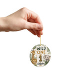 A hand holding a round personalized Christmas ornament against a solid white background. The ornament features a safari theme with illustrations of a giraffe, lion cub, elephant, and the text 'WILD ONE 1 Elijah 2026'.
