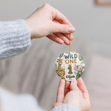 A person holds a round personalized safari-themed first birthday ornament with a gold hanging string. The ornament features illustrations of a lion, giraffe, elephant, and palm leaves, with the text 'WILD ONE', the number '1', and the name 'Elijah'.