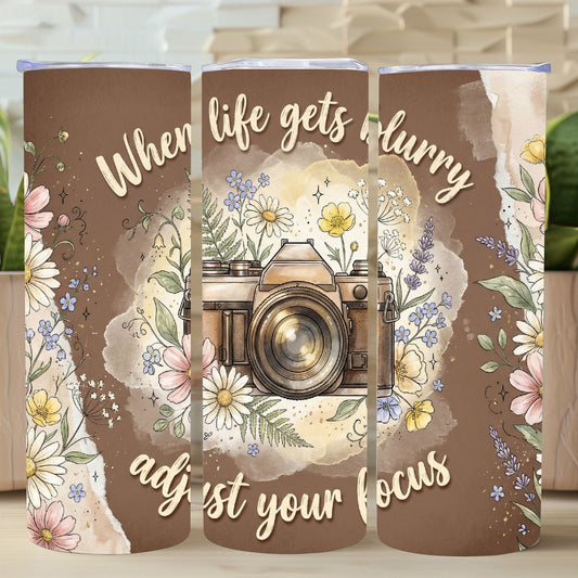 Tumbler with floral design and camera illustration with text 'When life gets blurry, adjust your focus' on a wooden surface.