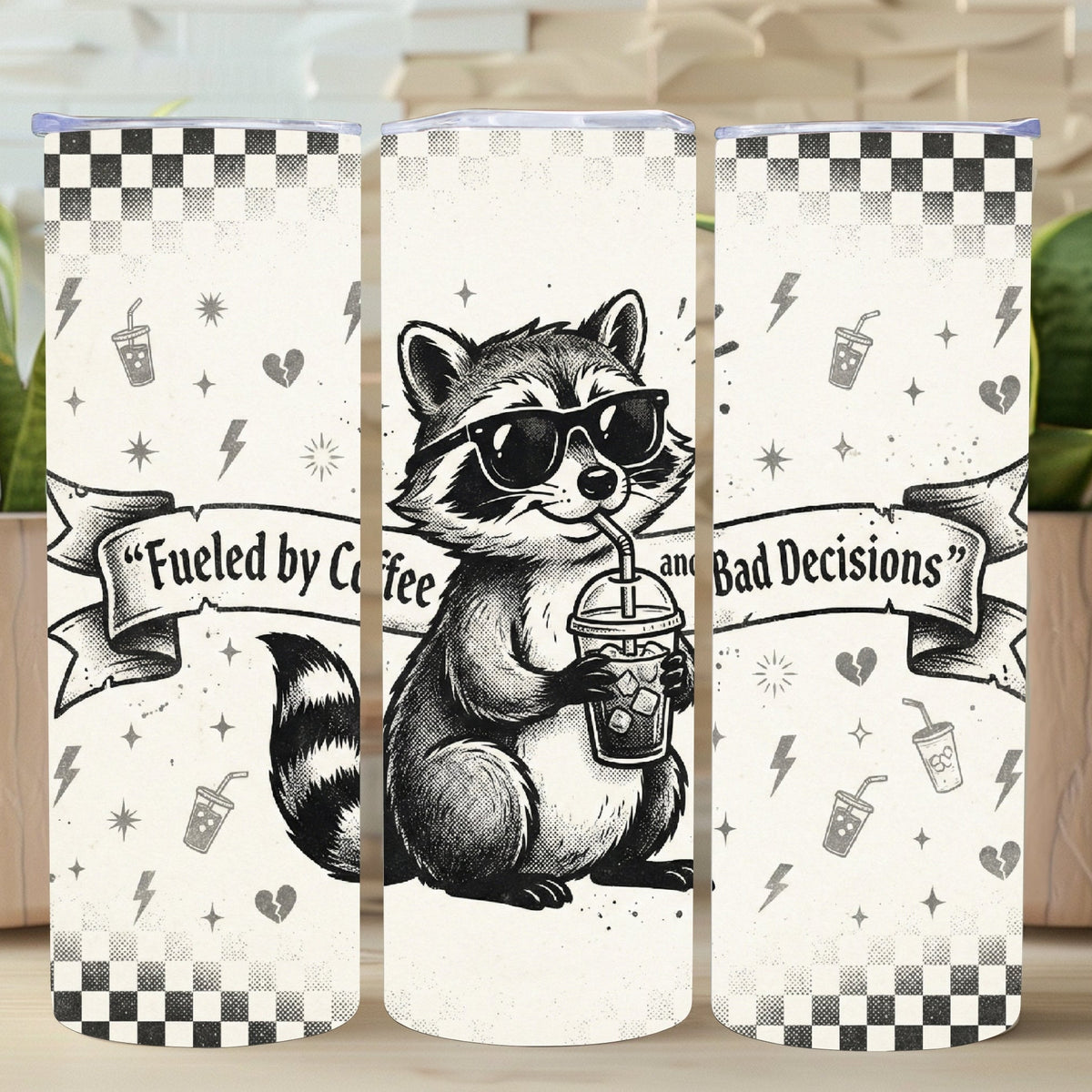 Three tumblers with a raccoon design and text on a wooden surface with plants in the background.