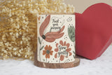 Decorative candle with floral design and motivational text, placed on a surface with dried flowers and a heart-shaped object in the background.