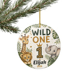 Round personalized Christmas ornament hanging from a gold string under pine branches. The ornament features safari animal illustrations including a giraffe, lion, elephant, and the text 'WILD ONE', a large number 1 with a crown, and the name 'Elijah' with '2026' below.